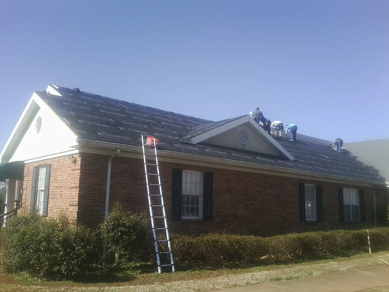 Crew tearing shingles off a brick ranch roof with a ladder set — Metal Roof Repair work by our crew in Bellport