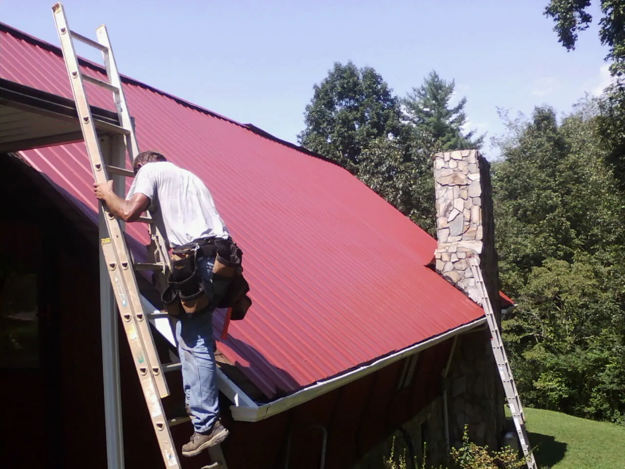 Roofer on a ladder beside a red metal roof and stone chimney — Soffit & Fascia Repair by Roofing Brothers in Bellport